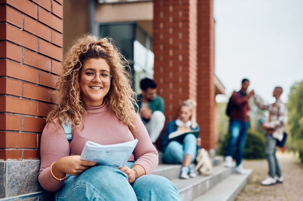 Happy female student learning while relaxing on steps at campus and looking at camera.